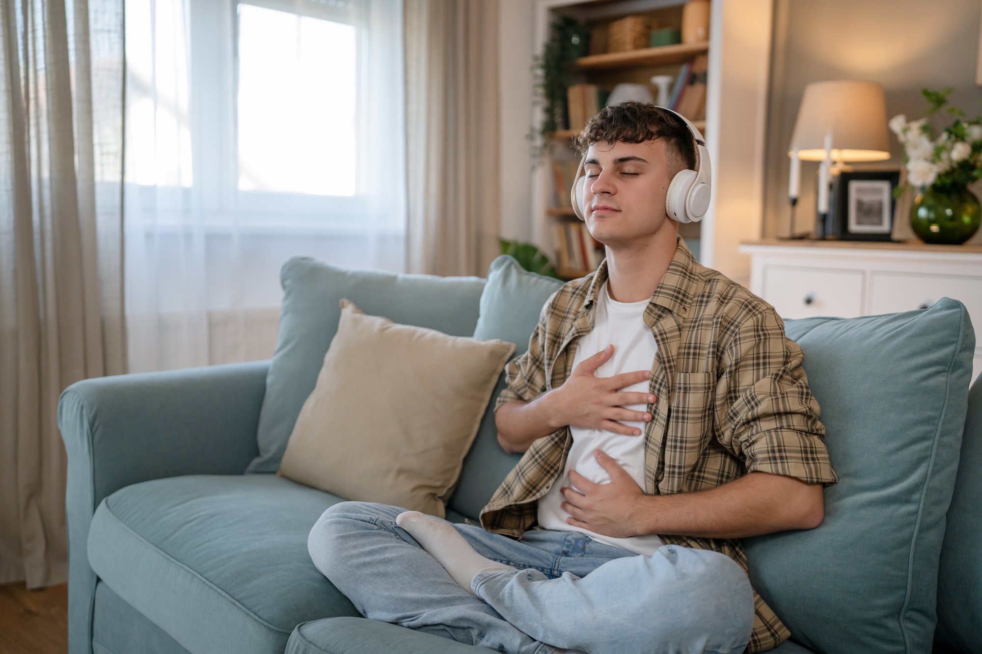 TN-Mental-Wellness-Trauma-young-man-sit-on-couch-eyes-closed-hands-on-chest-wearing-headphones-mediating